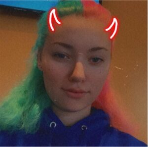 a girl with hair that is half aqua green and half peachy pink, pulled back from her face, smiles into the camera. a snapchat filter that looks like red neon devil horns is applied to her forehead. she is wearing a bright blue hoodie.