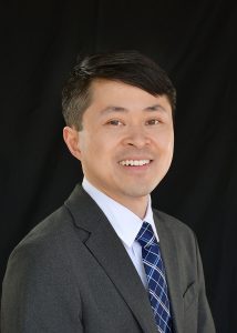 a professional portrait of Dr. Tong, who is wearing a dark suit jacket, a white formal dress shirt, and a blue with white plaid necktie. he is smiling at the camera and is in front of a dark studio backdrop.