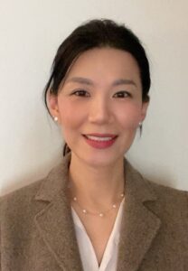 A portrait of Dr. Kim, who is smiling and standing in front of a white background. She's wearing a brown jacket over a white shirt.