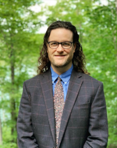 A photo of Dr. Randolph Ford, in front of a verdant backdrop. Dr. Ford is wearing a brown suit with a blue shirt and a brown floral tie. He has eyeglasses and wavy, shoulder-length dark hair.