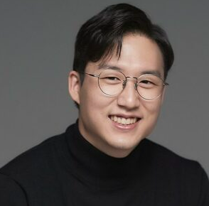 A formal portrait of Dr. Lee in front of a gray studio backdrop. He is wearing a black shirt and eyeglasses and is smiling.