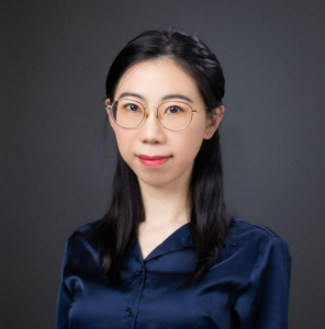 A portrait of Dr. Wang, taken in front of a gray studio backdrop. Dr. Wang is wearing eyeglasses and a dark blue button-down shirt. She has dark hair and is smiling. 