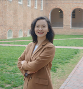 A portrait of Dr. Zhang, who is standing in the courtyard of a brick building. She is wearing a brown suit and has shoulder-length dark hair; she is smiling and her arms are crossed.