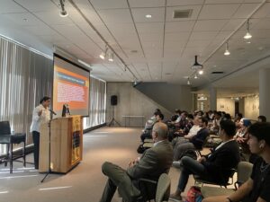 a view of many audience members at a lecture being given in a long, spotlit gallery space. a projection screen at the front of the room displays an orange powerpoint slide. 