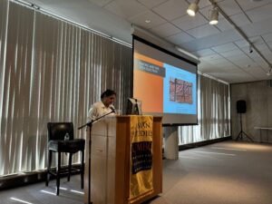 a lecturer stands at a podium in front of a wall of windows covered with vertical blinds. nearby, a projection screen shows an orange and white powerpoint slide.