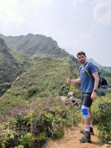 a man in a blue t-shirt, dark shorts, a backpack, and hiking boots stands on a dusty trail along the ridge of a mountain covered in scrubby green plants. he is turned to face the camera and is giving a thumbs-up gesture.