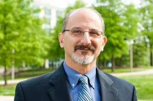 A portrait of Dr. Constantine Vaporis, a man with eyeglasses and a gray beard, standing in front of leafy green trees. He is wearing a blue shirt and necktie under a navy blue suit jacket.