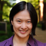 A portrait of Dr. Fan Yang, a woman with chin-length dark hair, standing outside on a sunny morning in front of leafy green trees and brick buildings. She is smiling and wearing a purple shirt.