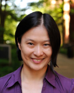 A portrait of Dr. Fan Yang, a woman with chin-length dark hair, standing outside on a sunny morning in front of leafy green trees and brick buildings. She is smiling and wearing a purple shirt.