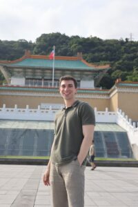 a man smiles for the camera as he stands in front of the National Palace Museum in Taipei, Taiwan on a sunny day.